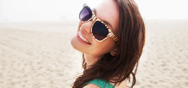 Woman in a green dress smiling over her shoulder at the beach, wearing sunglasses and looking back toward the camera
