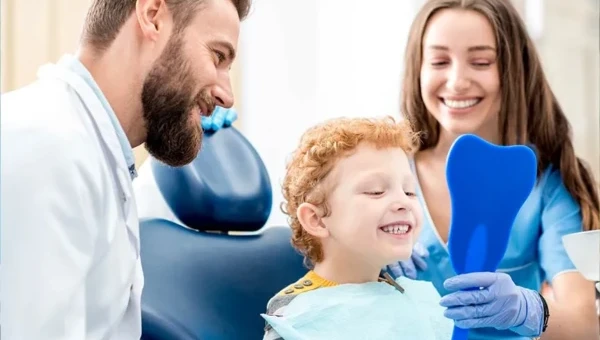 Smiling young boy sitting in a dental chair holding a blue mirror while dentist and hygienist look on happily.