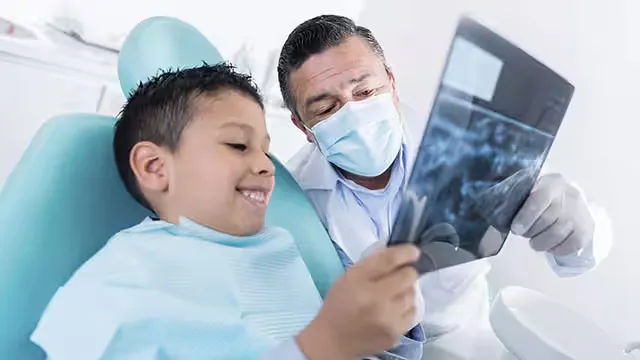 Smiling young boy in a dental chair holding and looking at a dental X-ray with a masked dentist beside him.