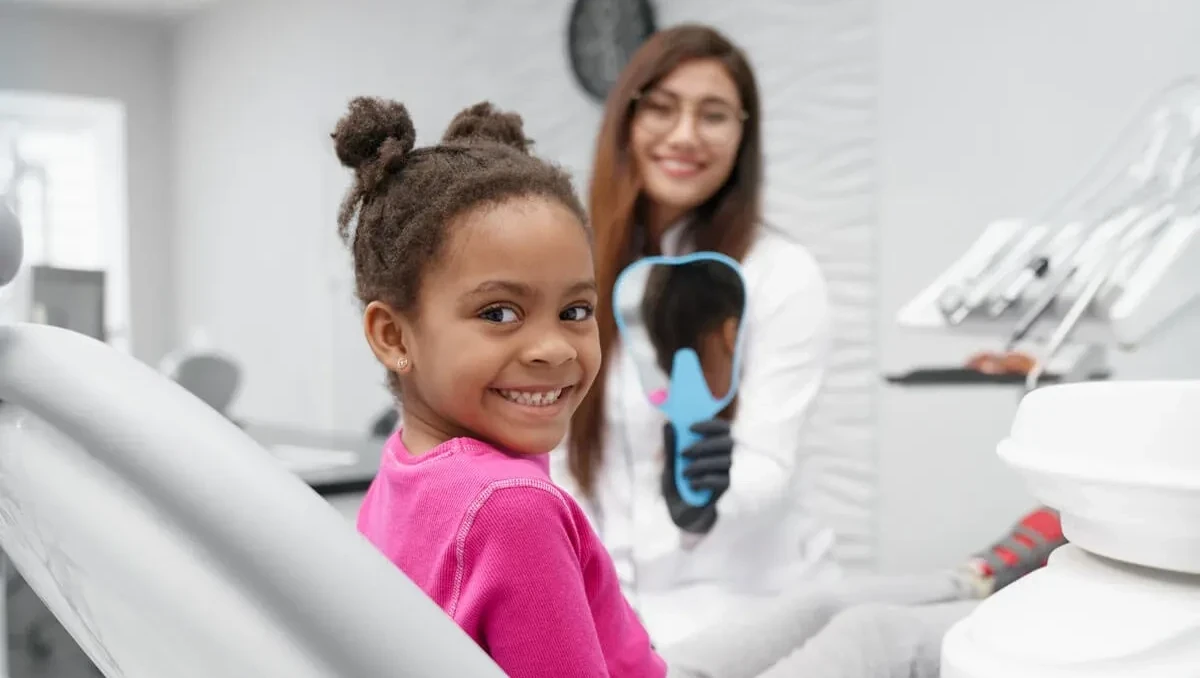 Young girl smiling in a dental chair while a dentist holds up a blue mirror behind her.