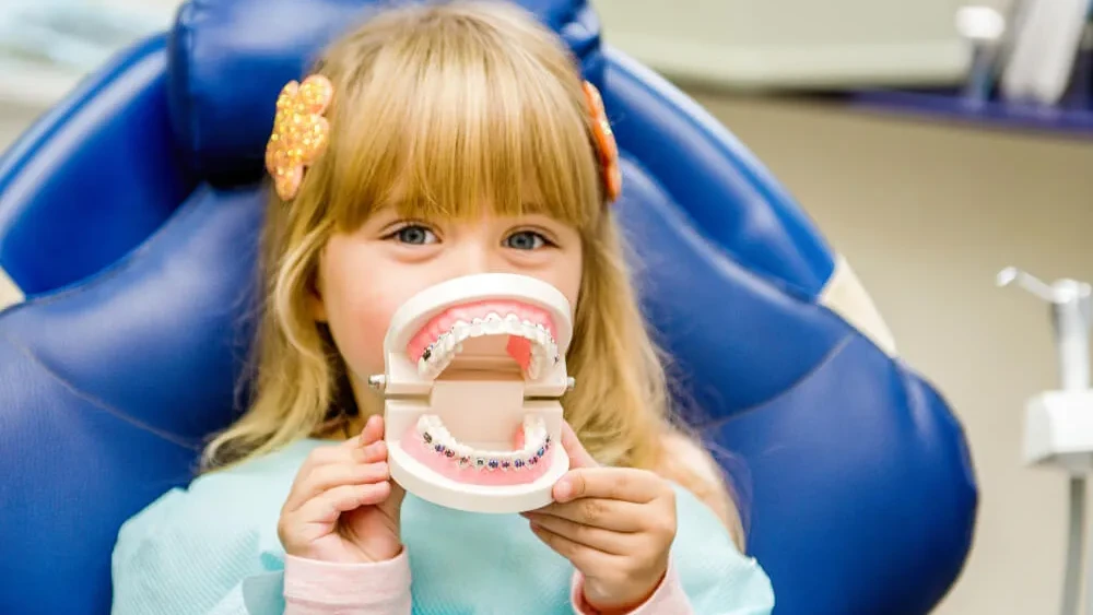 Little girl in a dental chair holding a model of teeth with colorful braces and smiling.
