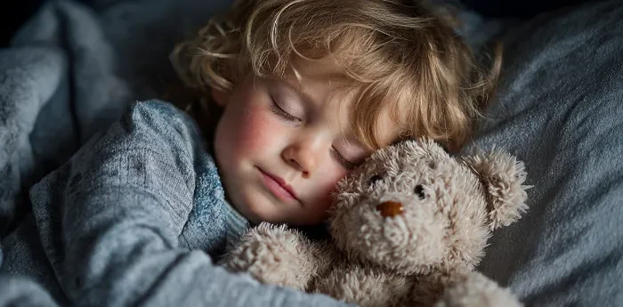 little boy in baby blue pajamas holding a teddy bear sleeping in bed.