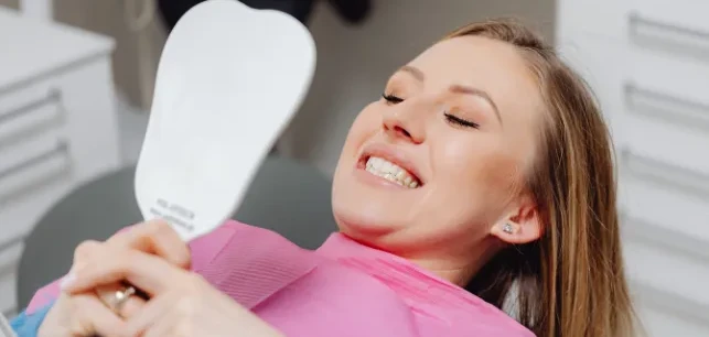 A woman laying in dentist chair with a pink dental napkin on, smiling into a white mirror she is holding.