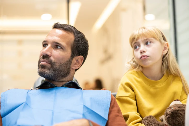 A man wearing a dental bib sits beside a young girl holding a teddy bear in a dentist's office, both appear to be waiting for a dental appointment.